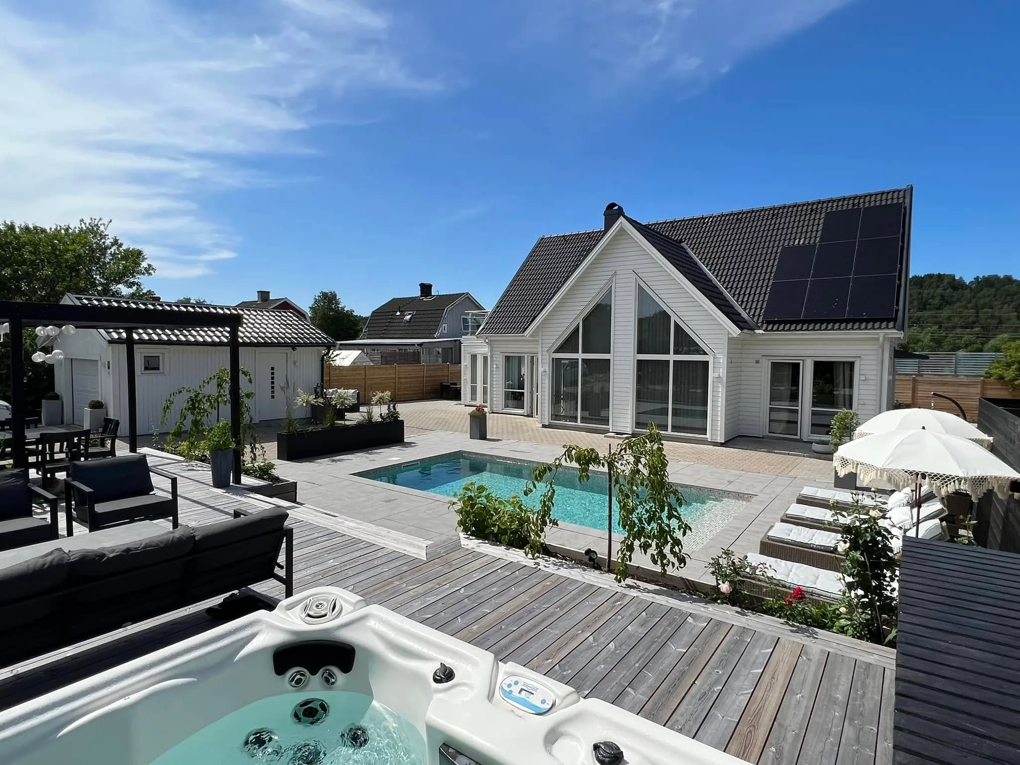 Poolaltan med utomhusspa, trädäck och solstolar framför modern vit villa