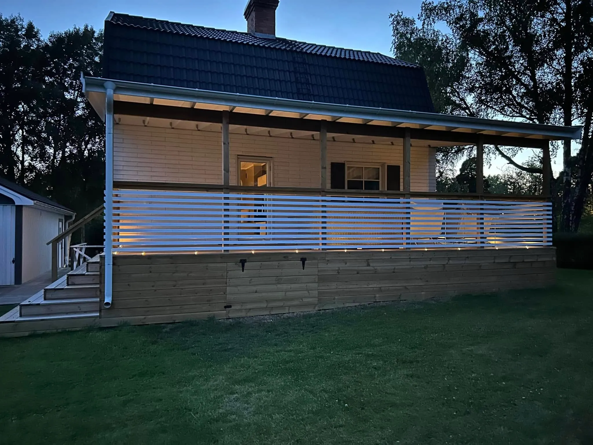 Upplyst altan med LED-belysning och horisontella vita räckesribbor runt ljust trähus i skymning