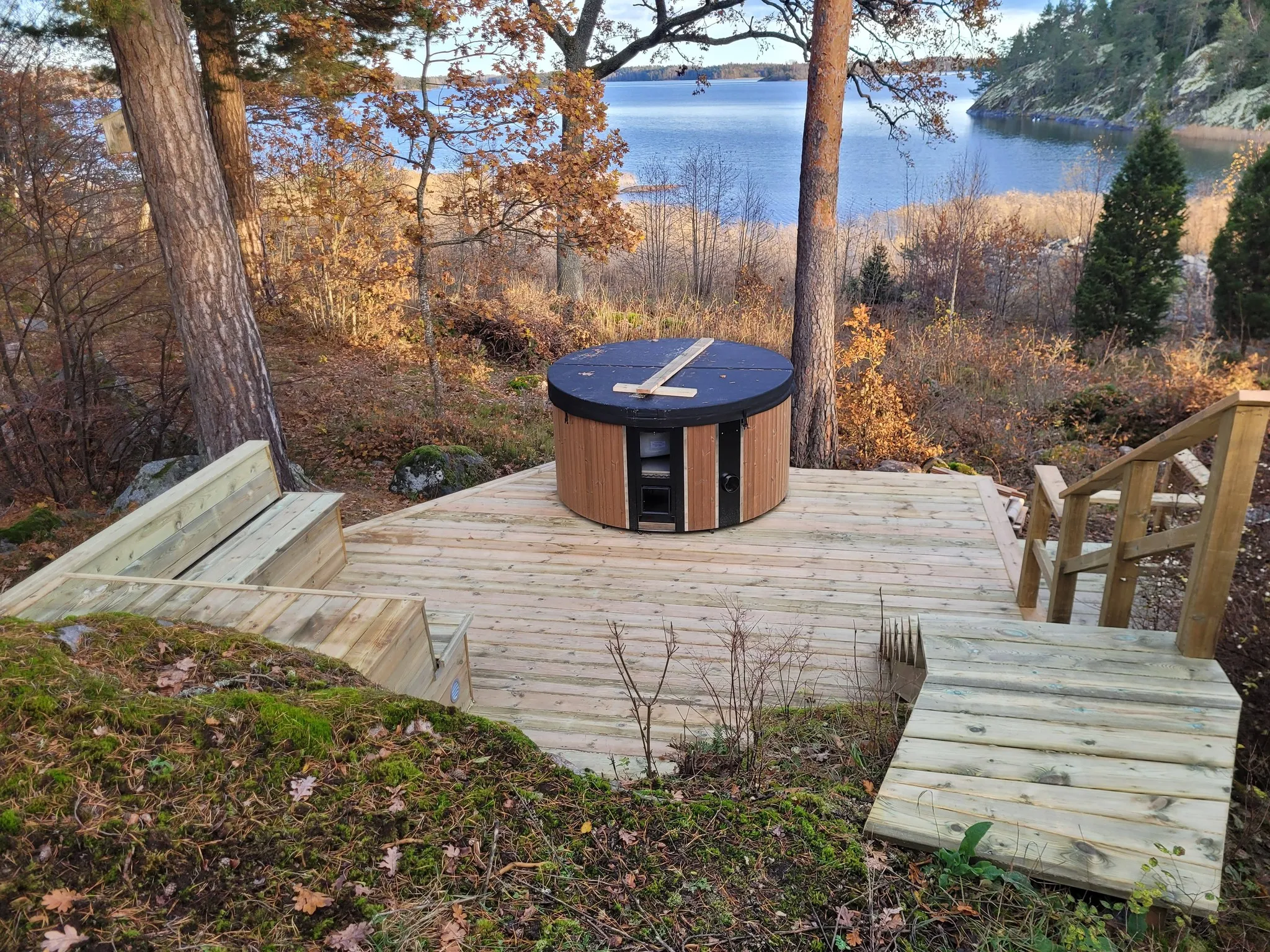 Litet trädäck med badtunna bland tallar och utsikt över sjö i höstfärger