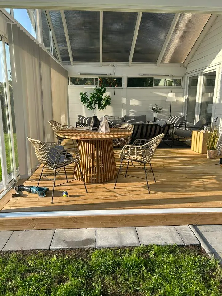 Rottingmöbler inne i ljust uterum med trädäck och grön trädgård